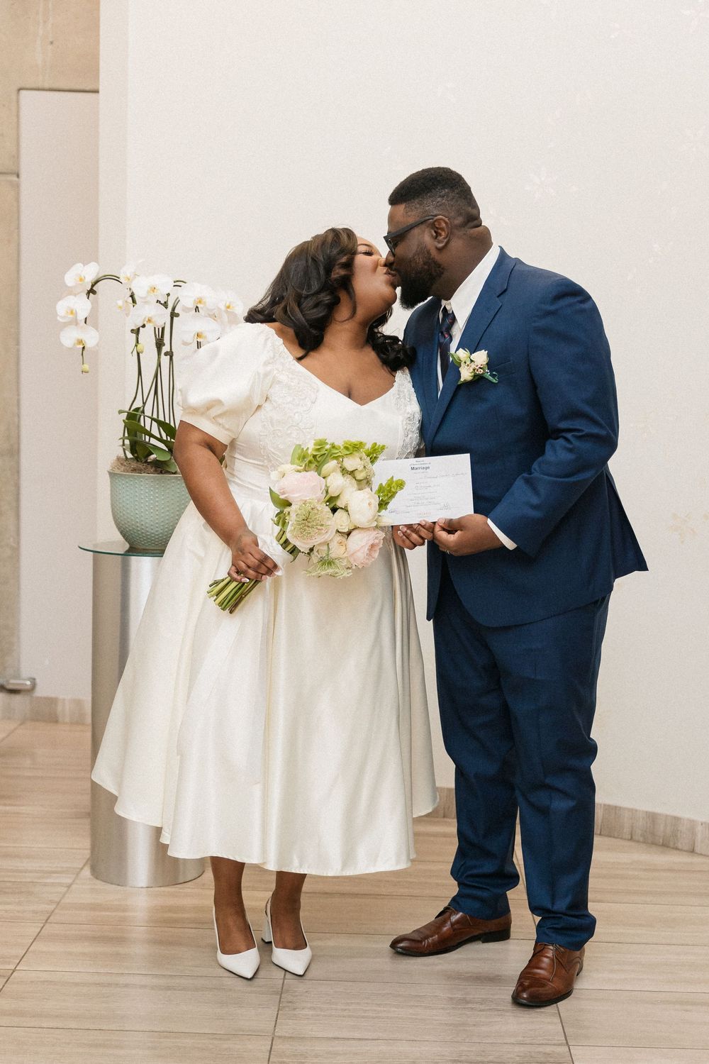 A couple shares an intimate moment at their wedding, with the bride holding white flowers and wearing a tea-length dress.