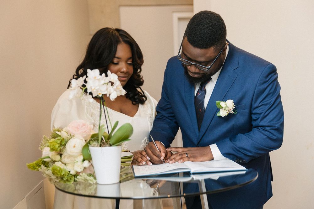 Newlyweds smiling while signing marriage documents beside white floral arrangements.