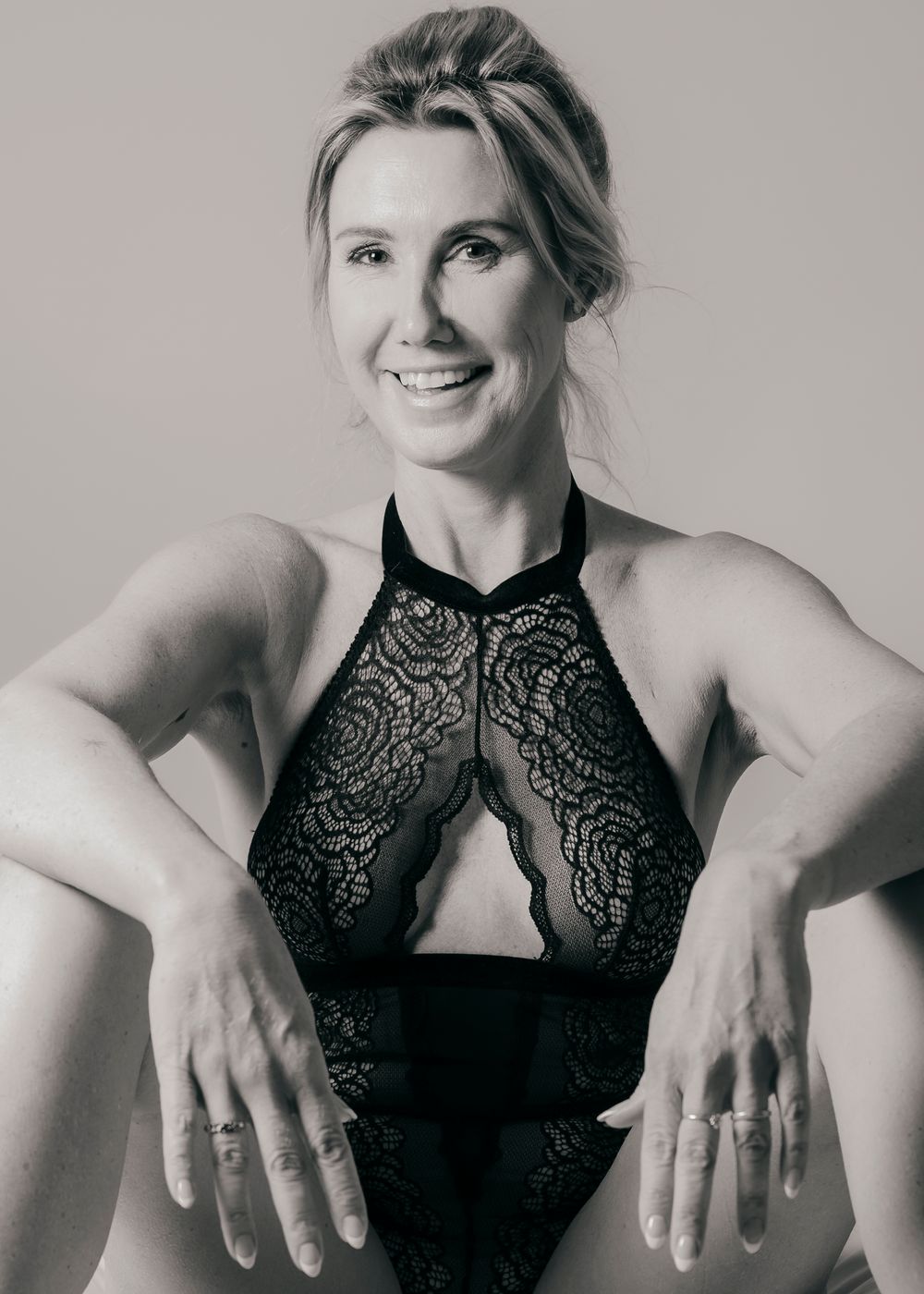Smiling blonde woman in black lace bodysuit in black and white studio portrait.