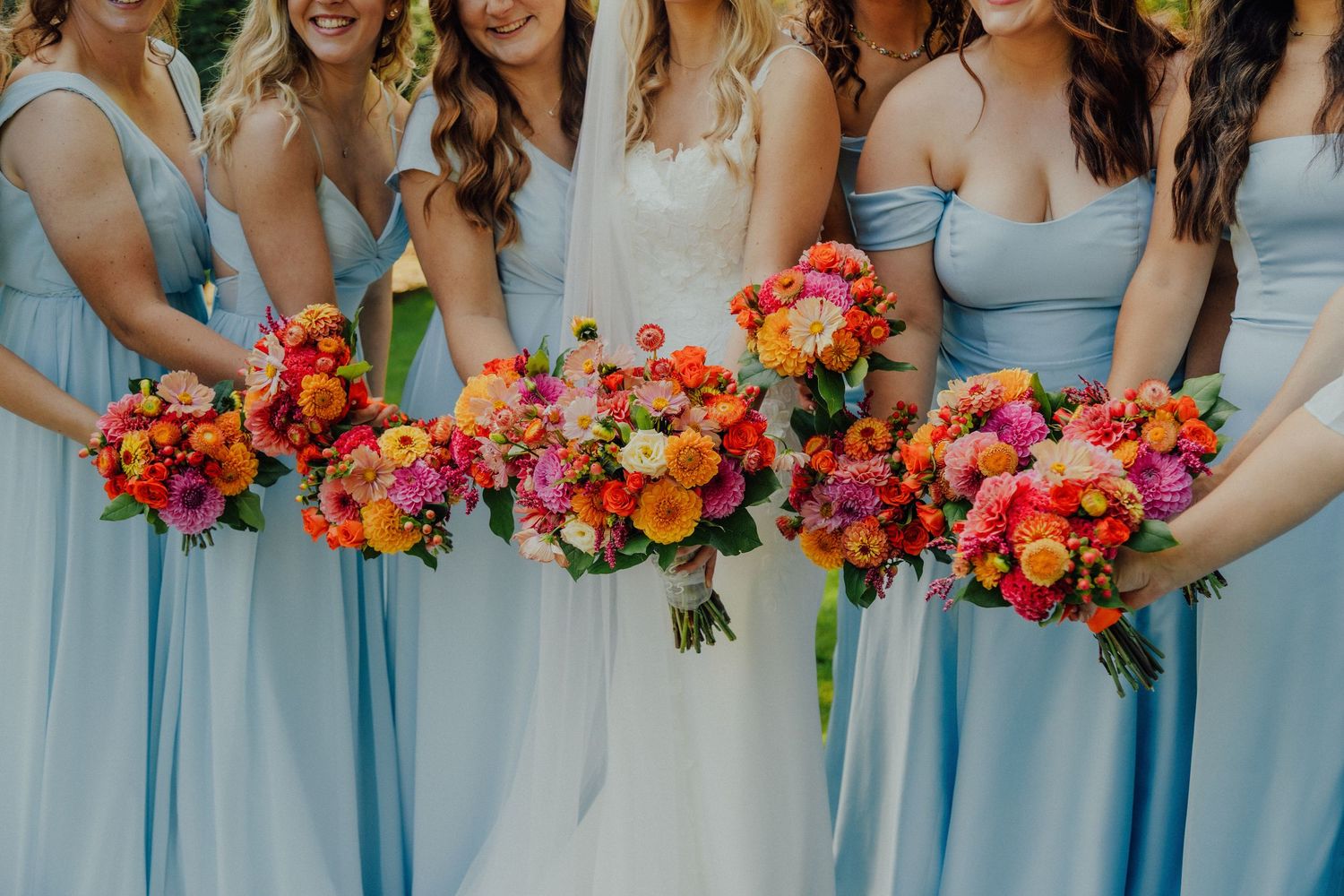 Bridesmaids in light blue dresses hold vibrant orange and pink bouquets while standing in a row outdoors.