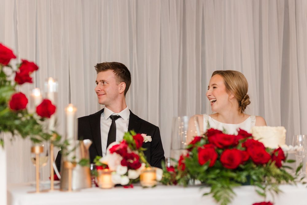 A wedding reception table decorated with red roses and white linens while guests enjoy dinner and conversation.