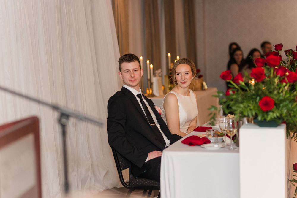 A couple sits at an elegant dinner table with red roses and white linens during a formal event.
