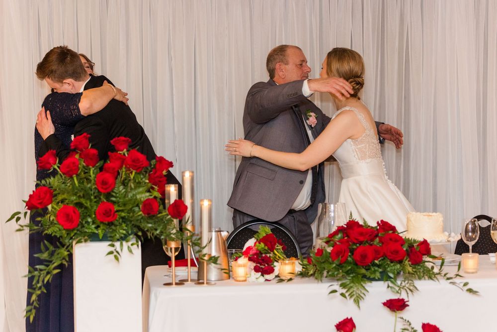 Wedding reception head table decorated with red rose centerpieces and white draping in an elegant banquet setting.
