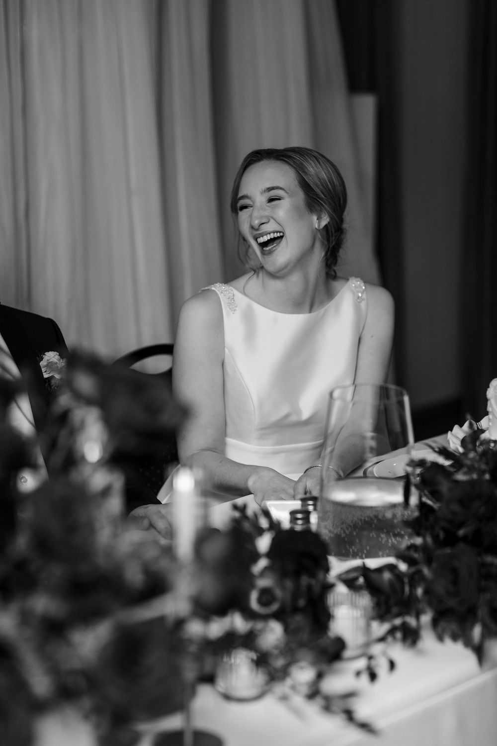 Black and white photo showing joyful reactions at an elegant wedding reception with floral centerpieces.
