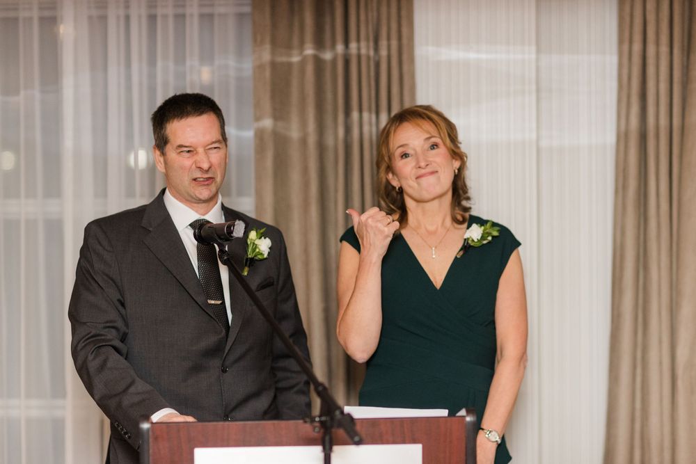 People in formal attire stand at a podium during a wedding ceremony in an elegant venue with curtained backdrop.