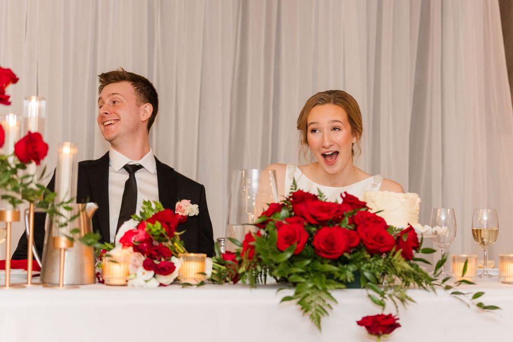 Newlyweds share a joyful moment at their reception head table decorated with red roses.