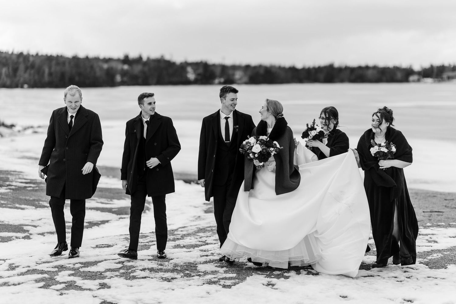 A wedding party in formal attire poses together on a snowy beach during winter.