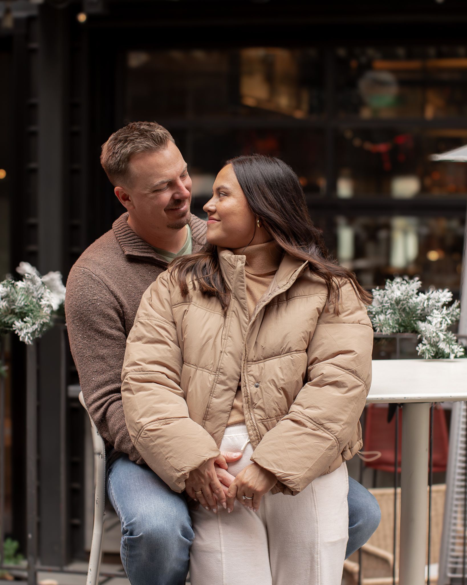 A couple in matching beige jackets share intimate moments while sitting outdoors near decorative white flowers.