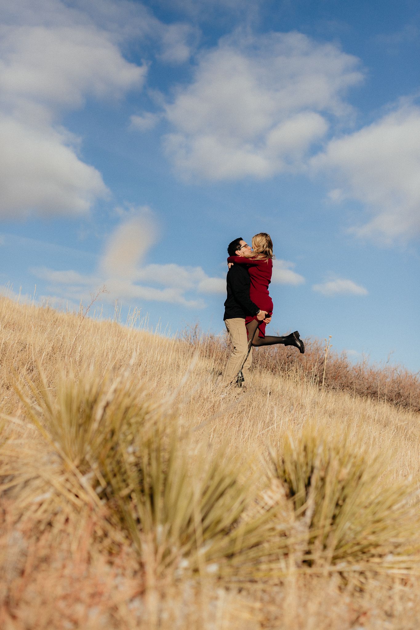 A couple embraces on a hilltop in a golden wheat field under a bright blue sky with white puffy clouds.