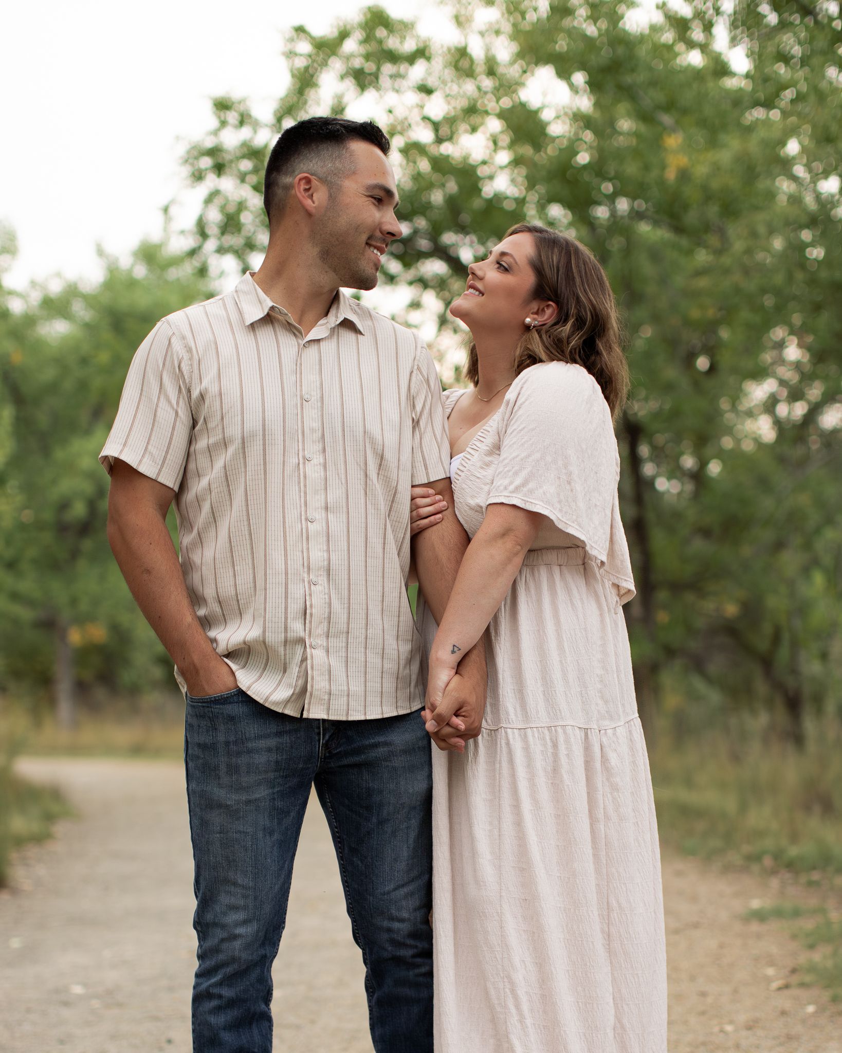 A couple shares a joyful moment together on a dirt path surrounded by green trees in casual summer attire.