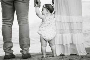 Black and white photo of a baby girl holding parents hands while learning to walk on a sandy beach.