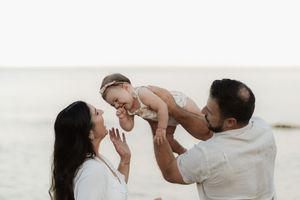 Happy family on beach with parents lifting laughing baby girl in the air near ocean shore.