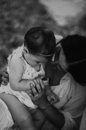 Black and white photo of a parent tenderly holding and gazing at a baby in a floral outfit outdoors.