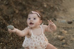 Adorable baby girl in floral romper and pink bow headband sitting outdoors, reaching out with a curious expression.