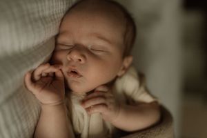 Peaceful newborn baby sleeping with tiny hand raised to cheek, soft natural light, close-up portrait photography.
