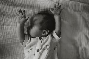 Black and white photo of a newborn baby lying on back with arms raised, wearing a white onesie.