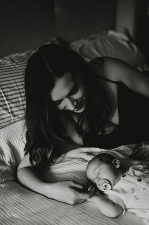 Black and white photo of a mother tenderly gazing at her newborn baby lying on a bed with a pacifier.