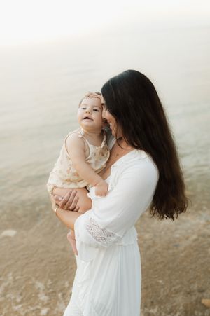 A mother in a white dress holds her smiling baby girl near the water's edge in a serene outdoor setting.