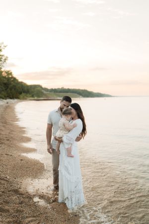A couple stands on a sandy beach at sunset, holding their baby, dressed in white and neutral tones with calm water behind.