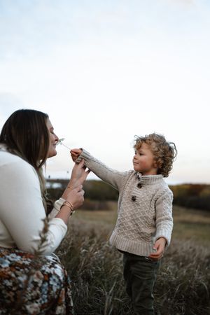 A child in a knit sweater reaches out while standing in a field at sunset with someone kneeling nearby at family lifestyle session