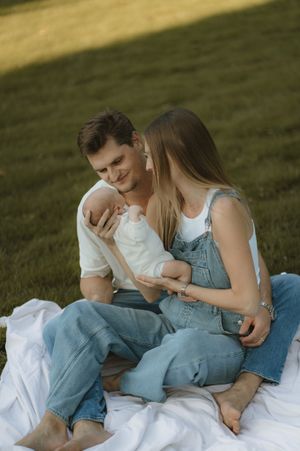 A couple sits on a white blanket in grass, smiling at their newborn baby, both wearing casual denim outfits.