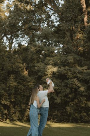 A joyful couple in matching white tops and jeans lift their newborn baby in a sunlit green park.