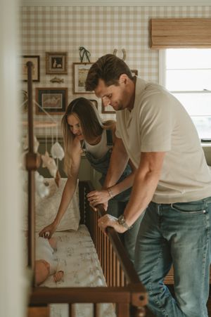 A couple lovingly looks down at their newborn baby in a wooden crib in a charming nursery with plaid wallpaper.