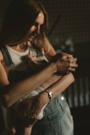A woman in overalls tenderly holds hands with an elderly person, wearing a gold bracelet, in warm moody lighting.