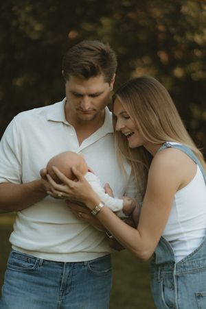 A couple in jeans and white tops lovingly cradle their newborn baby outdoors in a lush green park setting.