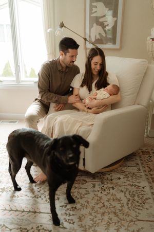 A couple admires their newborn baby in a white rocking chair while a black Labrador stands nearby in a bright nursery.