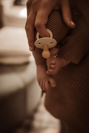 Close-up of adult hands cradling tiny newborn baby feet with a pacifier, wearing a cozy knit sweater.