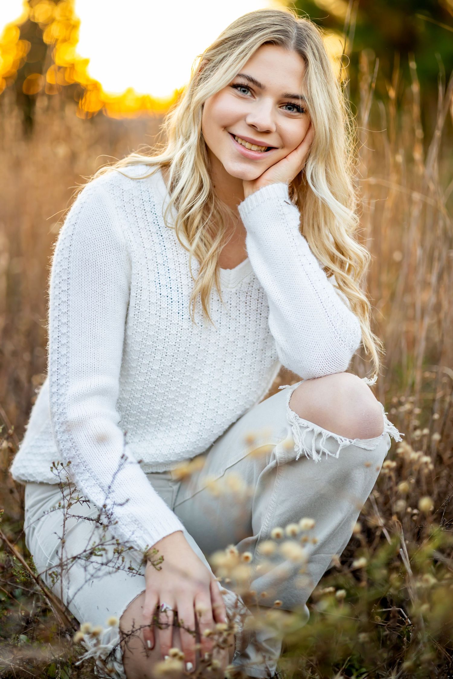 A person in a white sweater poses in a golden autumn field during sunset while holding their hair.