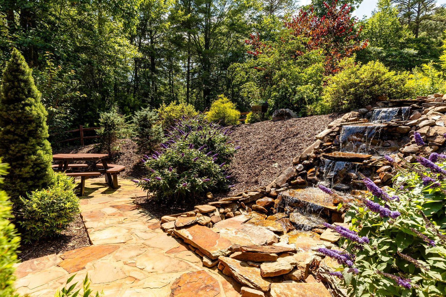 A waterfall feature surrounded by lush landscaping and a picnic table on the stone patio.