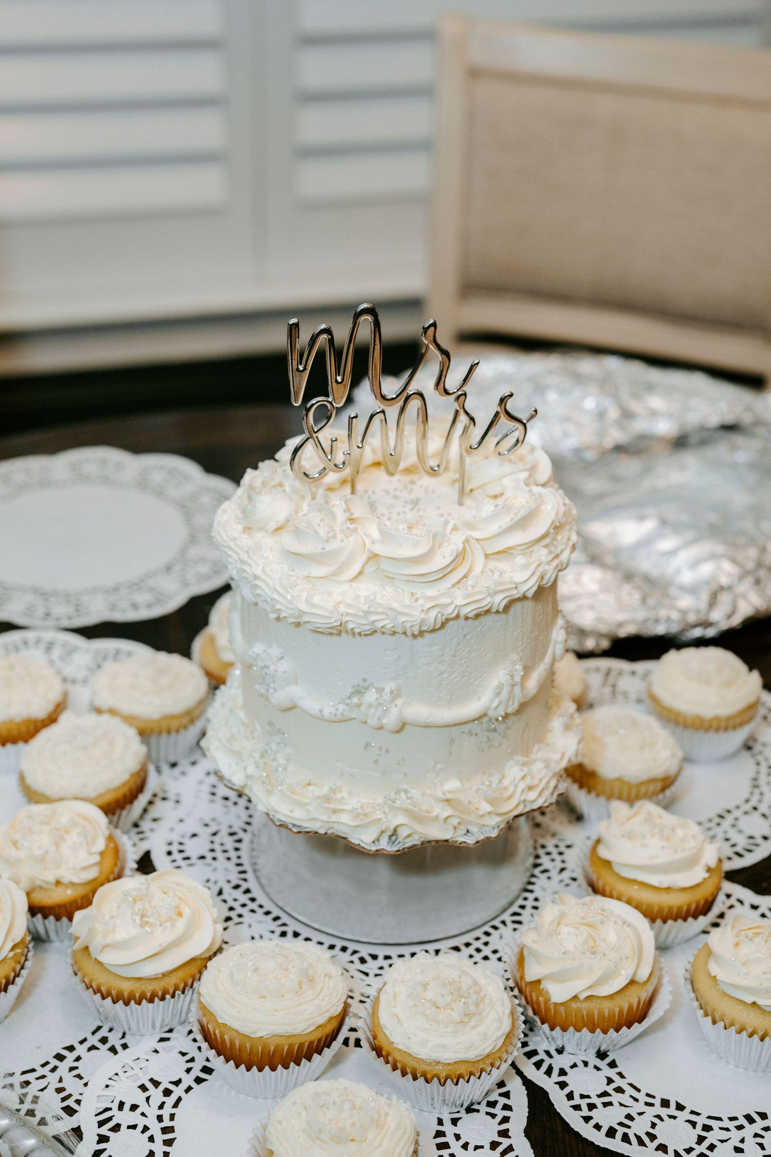 A series of white wedding cupcakes arranged around a small white frosted cake with a gold topper on a lace doily.