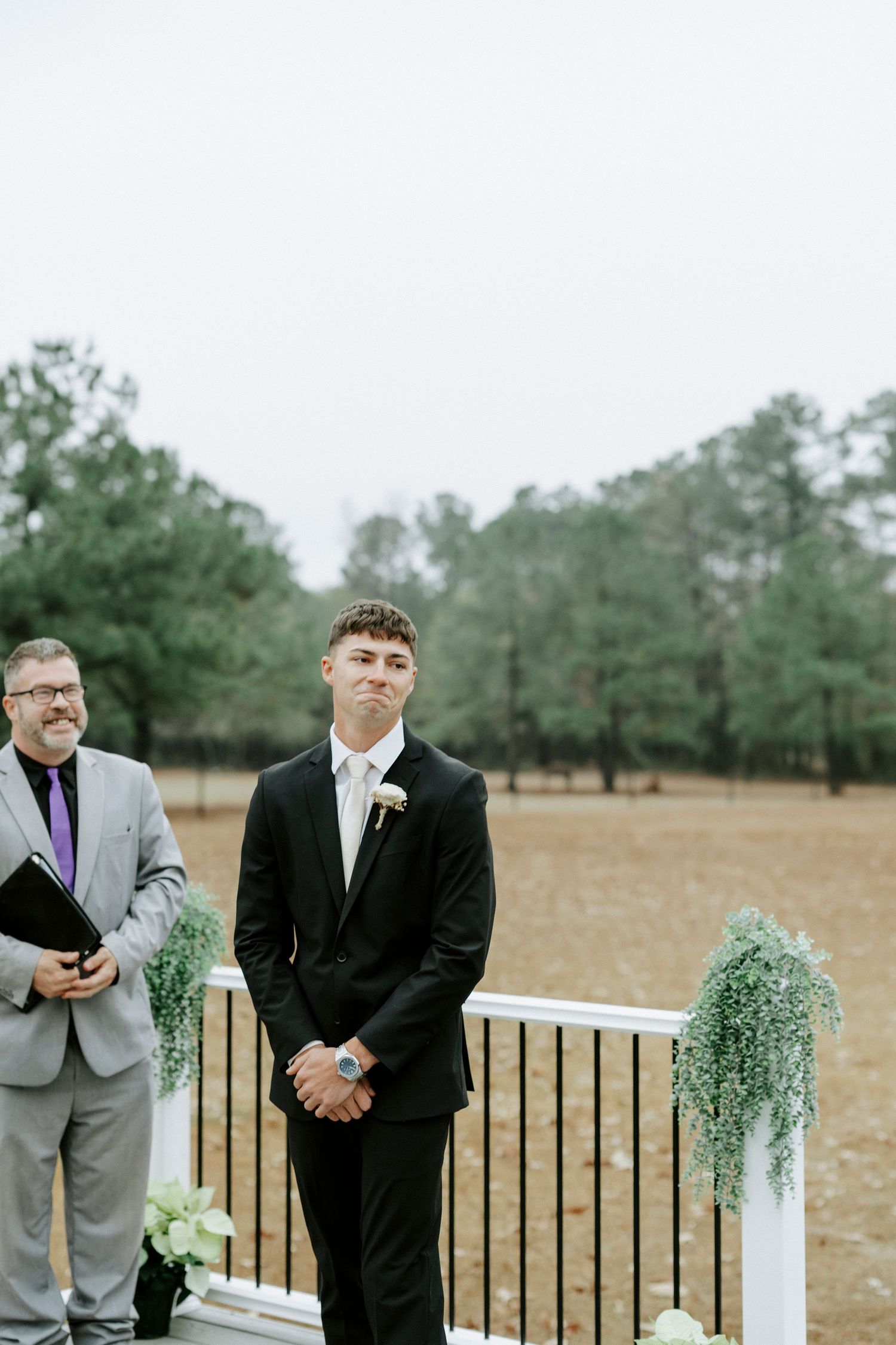 Groomsmen in black and gray suits share emotional reactions during an outdoor wedding ceremony.