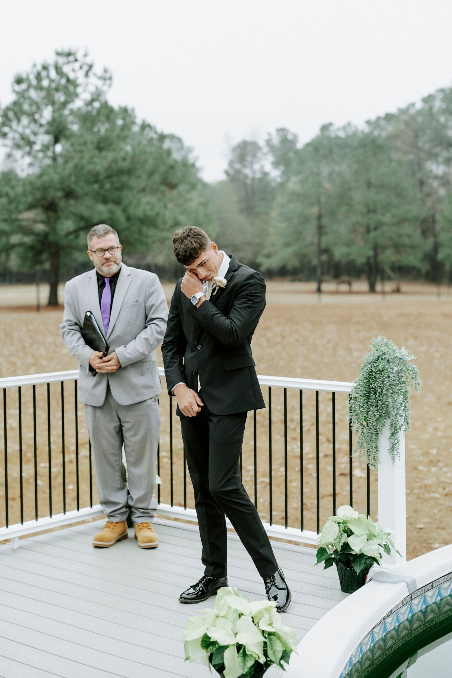 Two people in formal attire lean over a wooden deck railing to look at something below at an outdoor venue.