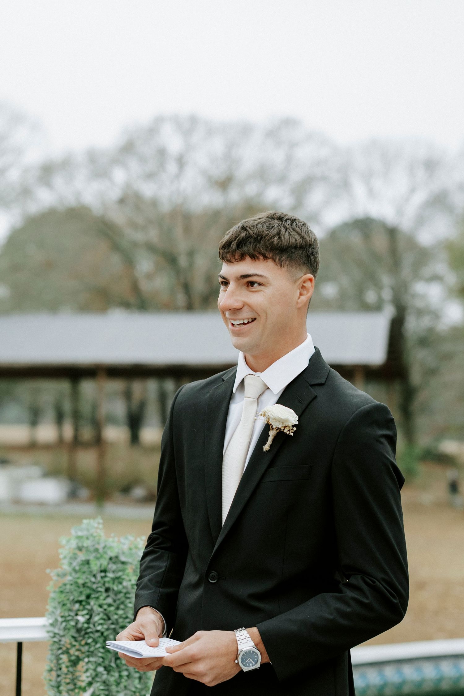 A person in a black tuxedo and white tie smiling while holding a small potted plant outdoors.