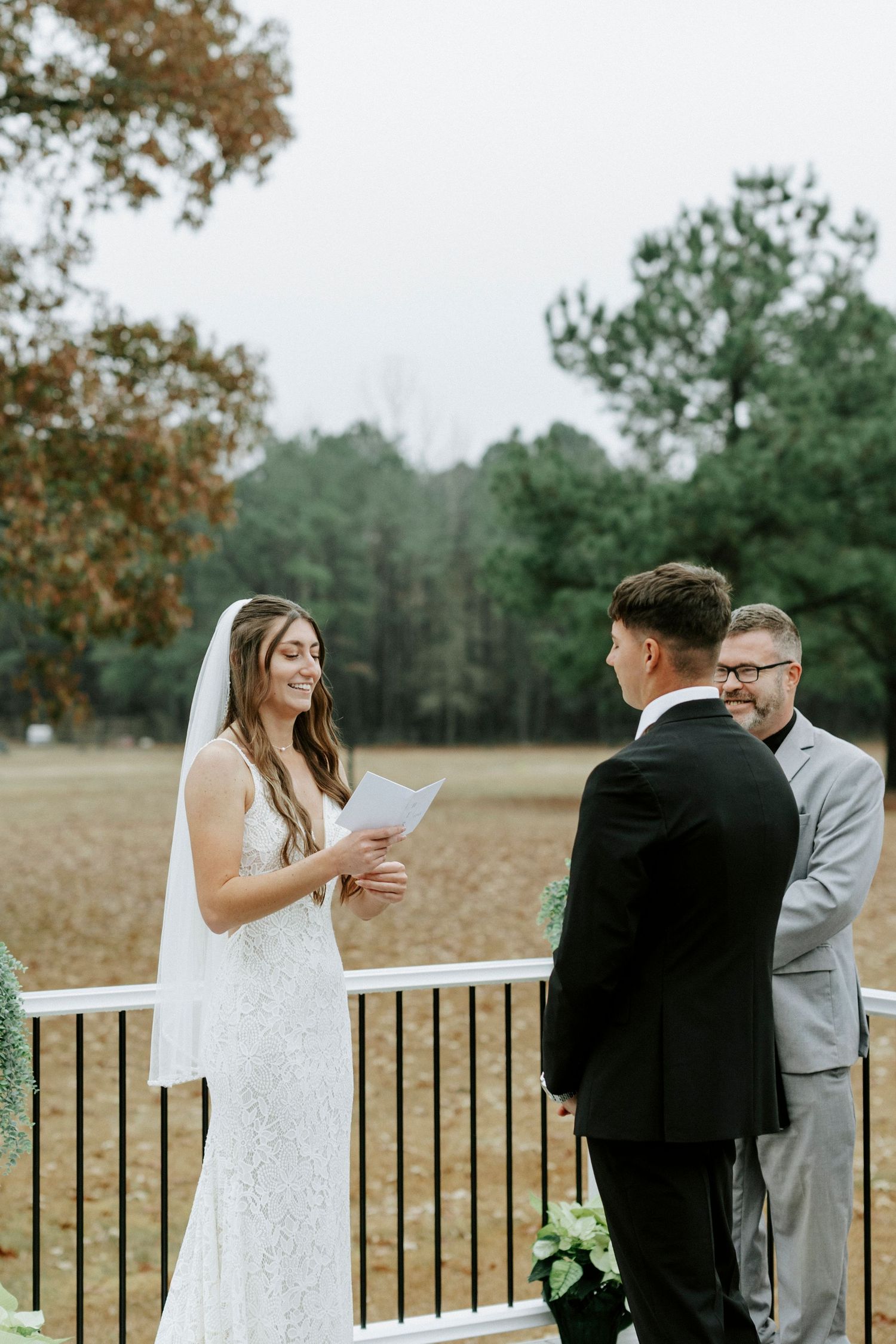 A couple exchanges vows during an outdoor wedding ceremony with a wooded backdrop and autumn foliage.