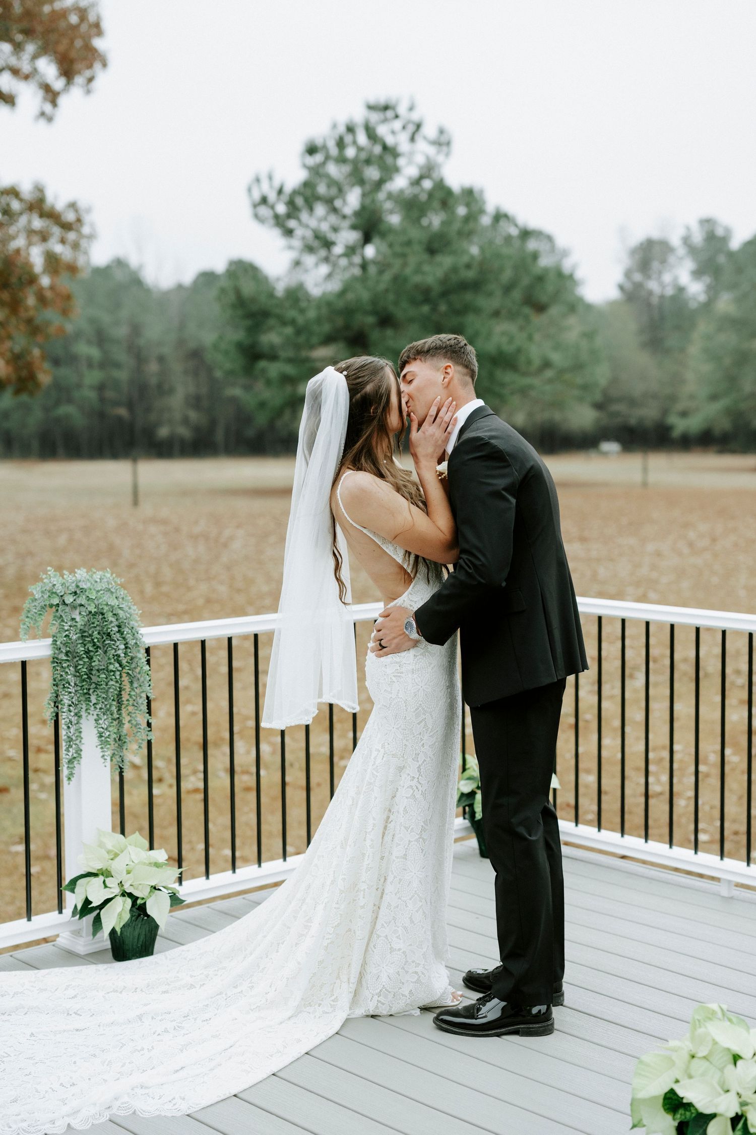 Newlyweds share a romantic kiss on a white wooden deck with a long flowing veil and autumn scenery in the background.