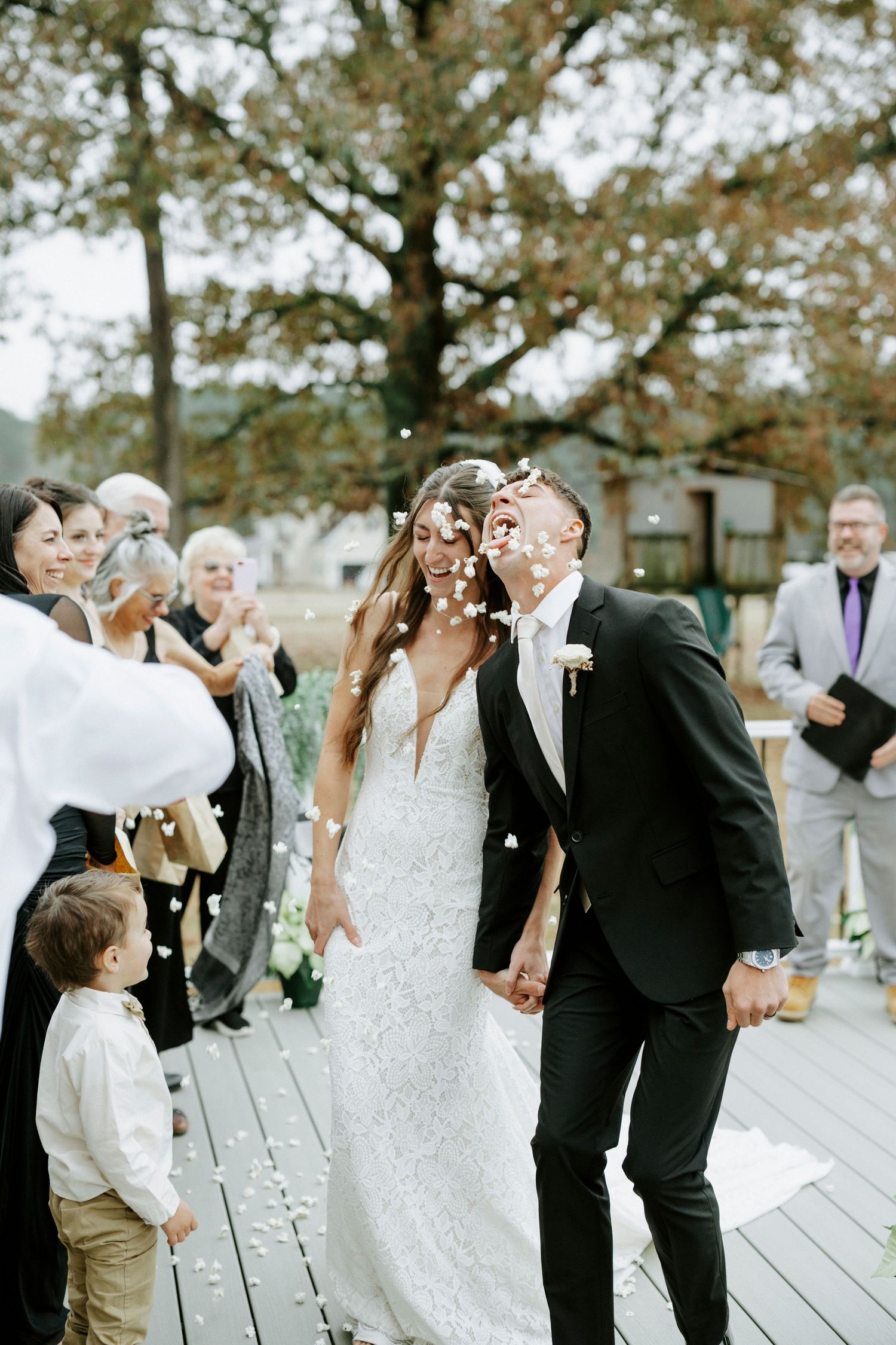 Newlyweds celebrate with guests on a wooden deck as confetti falls during an outdoor wedding ceremony.