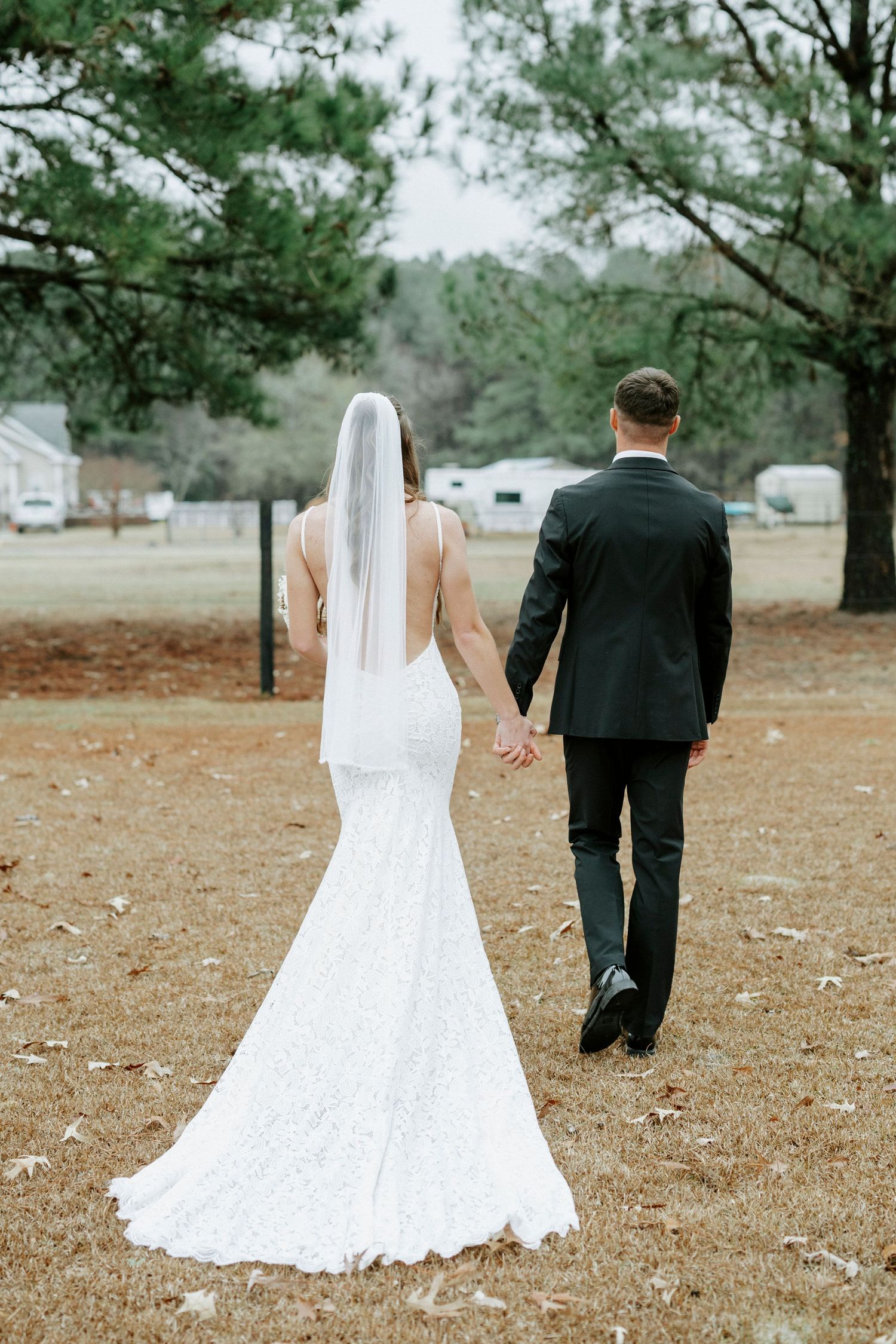 A bride in a fitted lace gown and groom in black suit walk hand-in-hand through a rustic outdoor setting.