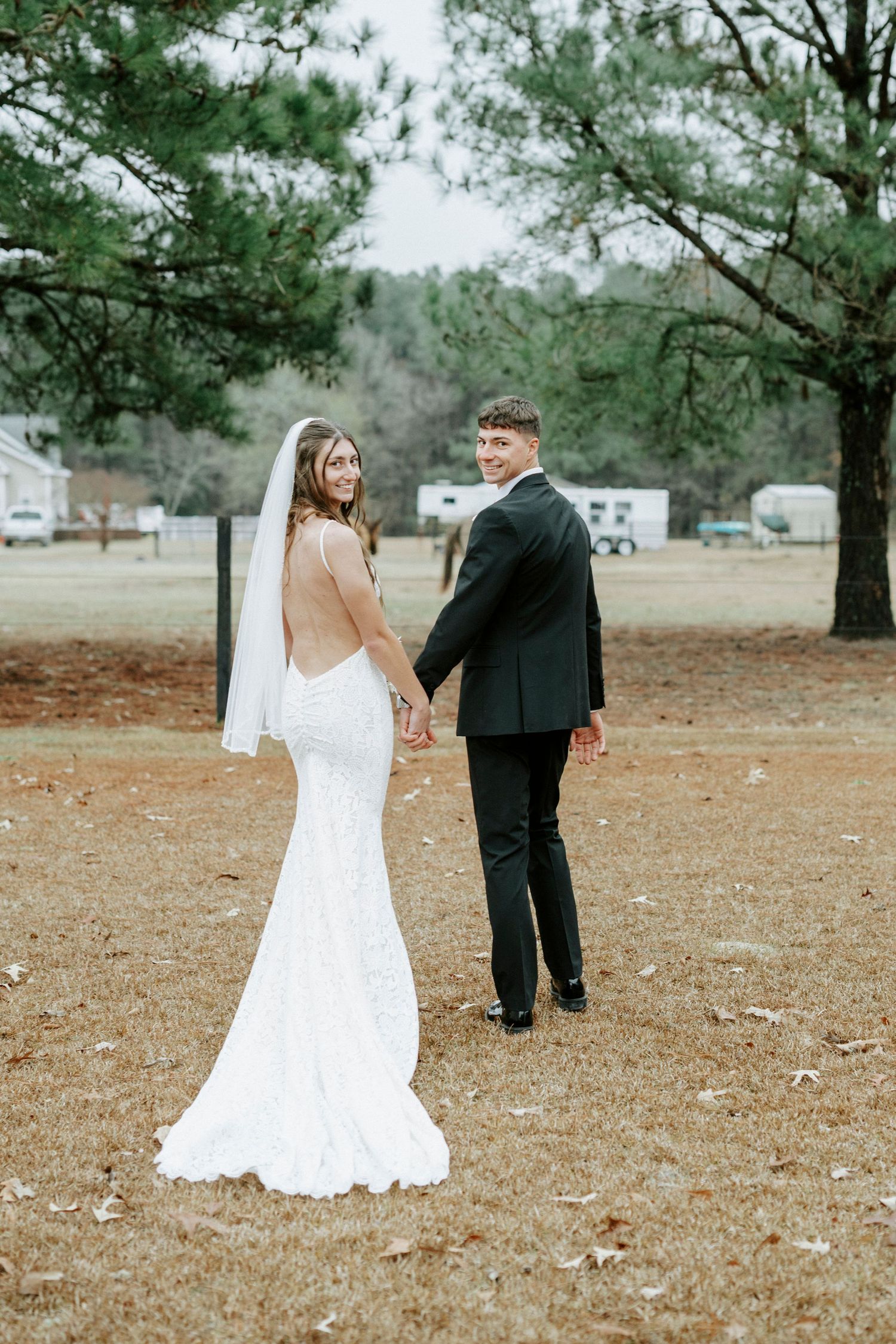 A bride and groom in formal wedding attire walk together on fallen leaves in a rustic outdoor setting.