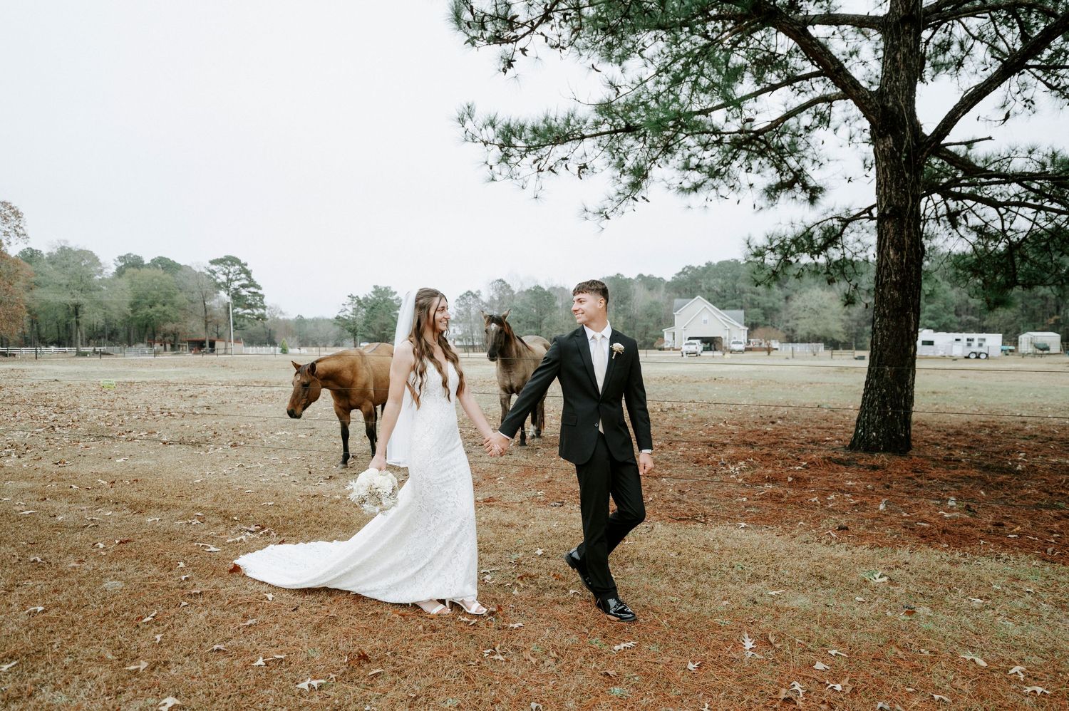 Couple in wedding attire walks through a scenic outdoor setting with tall trees and autumn foliage.