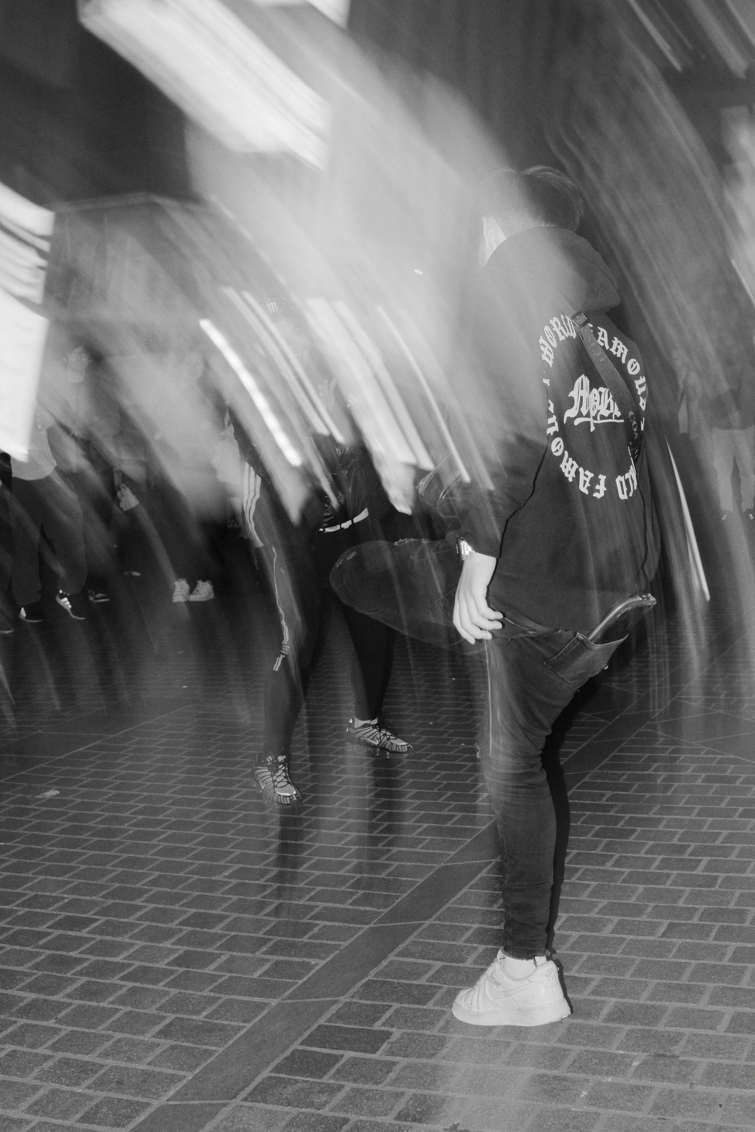 Black and white long exposure photograph showing motion blur of dancers performing on a tiled floor.