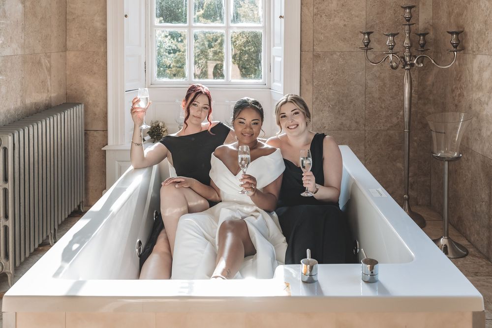 A relaxed photo of bride and bridesmaids celebrating her wedding day in a jacuzzi bath with a glass of fizz.