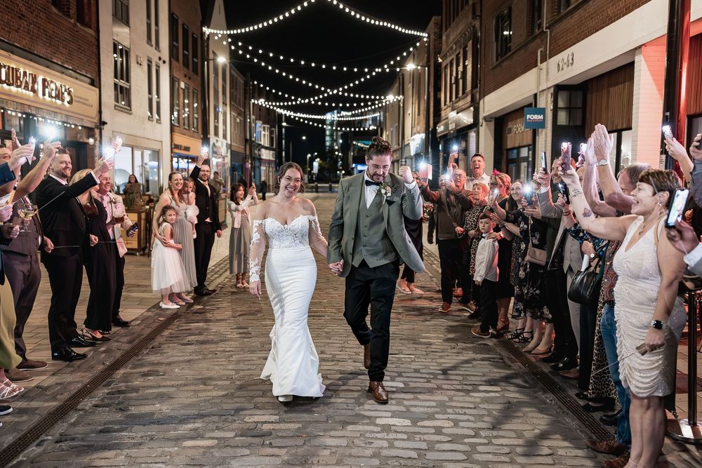 Wedding party walks through a charming cobblestone street lined with guests holding mobile phone lights at night.