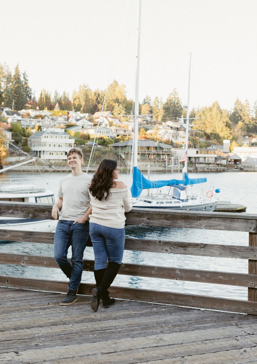 A couple sits together on a wooden dock overlooking a marina with boats and houses in the background at sunset.
