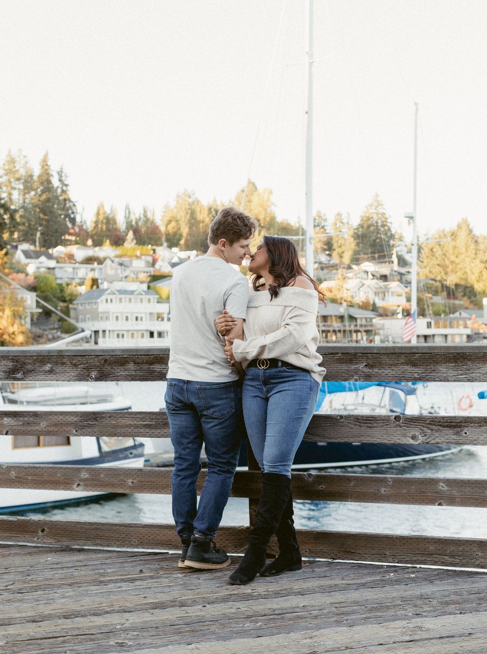 A couple shares a romantic moment on a wooden dock overlooking a marina with autumn trees in the background.