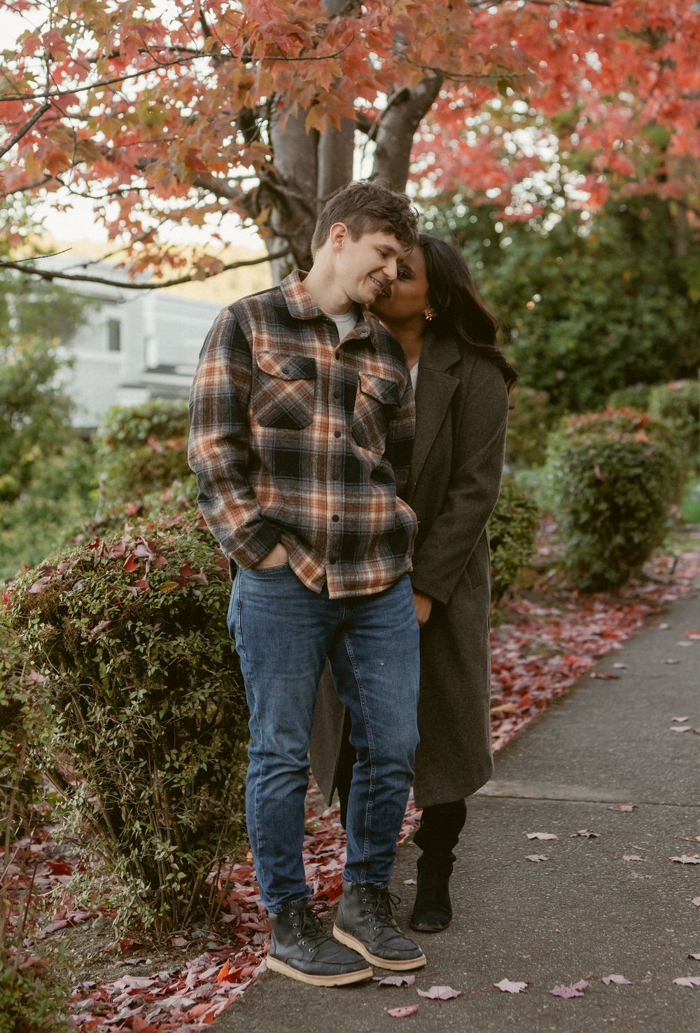 Couple sharing a tender moment together on an autumn sidewalk with red maple leaves falling in the background.