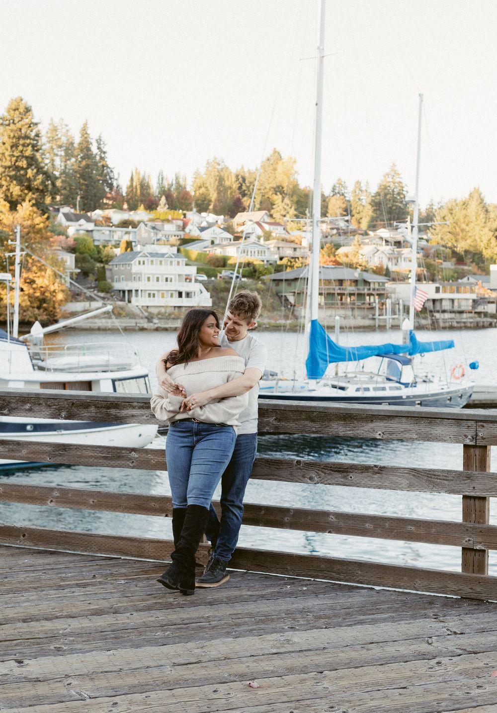 A couple shares a romantic moment on a wooden dock overlooking a marina with boats and autumn trees in the background.